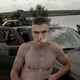 Ukrainian soldiers rest by a car; a shirtless man wearing a rosary stares at the camera while exhaling smoke.