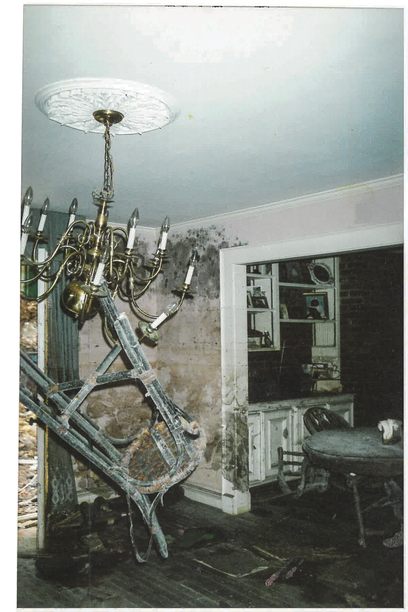 photo of house interior lit by camera flash with twisted metal hanging from chandelier, mold on walls, and large wood dining table just visible in next room