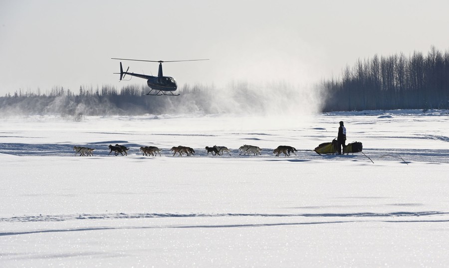 A helicopter flies near a sled dog team as it travels on a frozen snow-covered river.