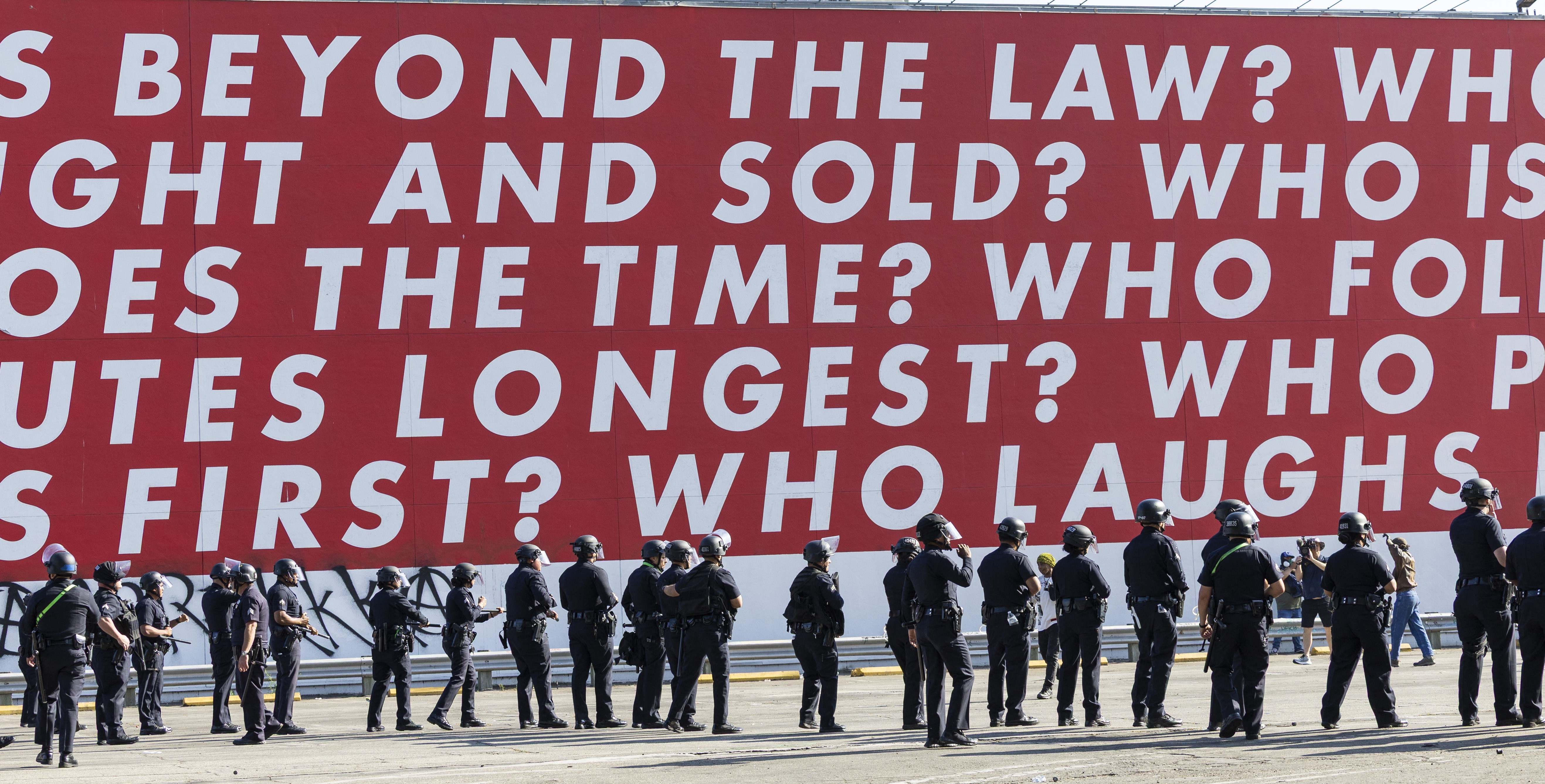 A line of riot police officers stand beneath a huge work of art on an outside wall, made up of text, reading in part 