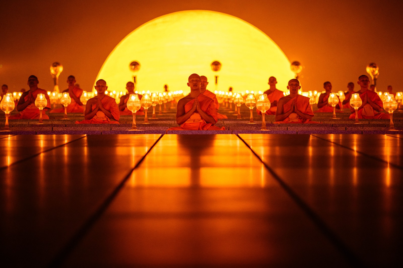 Buddhist monks sit in prayer, arranged in rows in front of a glowing golden dome.