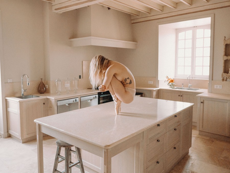 A partially clothed woman wearing ballet shoes poses, crouching while on pointe, atop a kitchen counter.