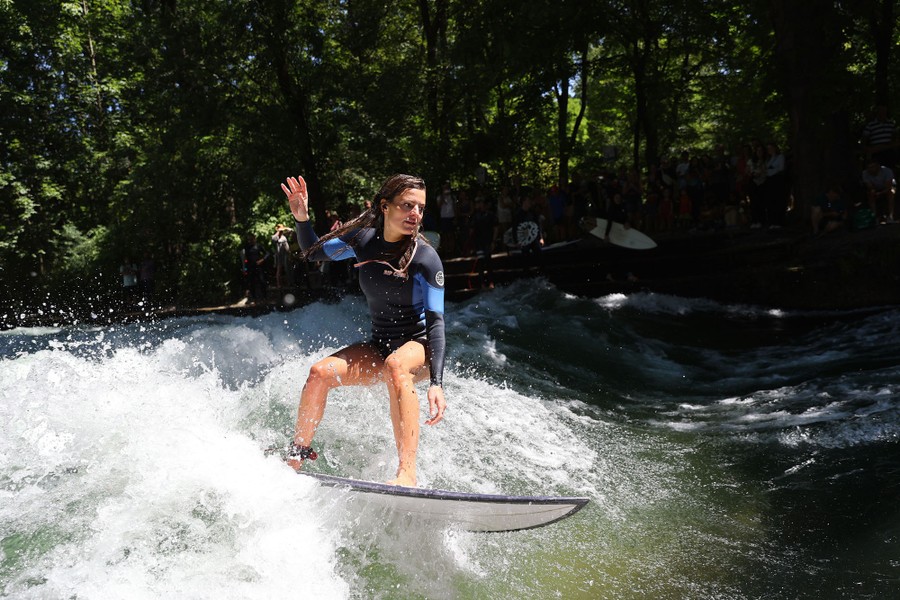 A person rides a surfboard on a small wave in a river in a park.