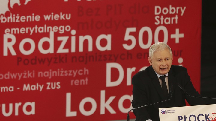 Jaroslaw Kaczyński, the leader of Poland's governing party, stands at a lectern in front of a banner with his party's policies.