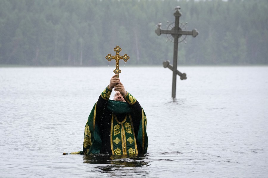 An Orthodox priest stands in a lake, holding a gold cross above his head. A silver cross stands behind him in water.