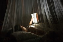 A girl reads a book at night under mosquito netting.