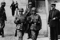 Black-and-white photo of four Nazi soldiers walking through a city street with rifles strapped to their backs as a policeman watches