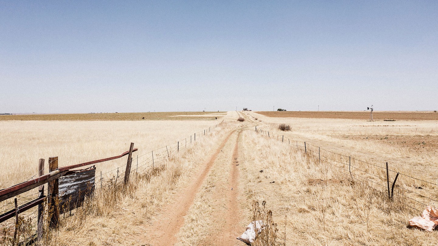 A color photo looking down a dirt road lined by wire fencing and surrounded by dry fields.
