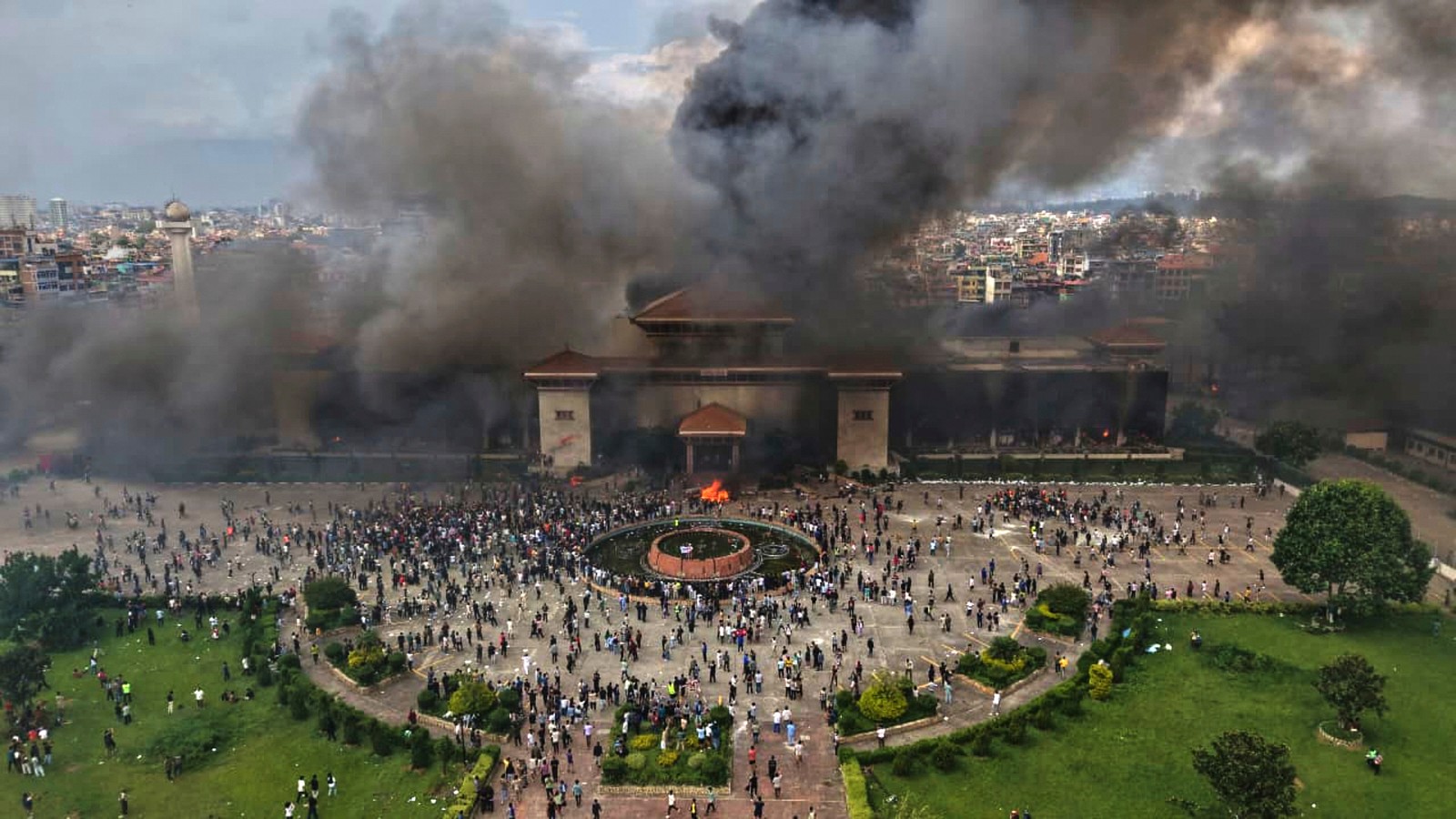 An aerial view of a large government building with dark smoke rising from it, as demonstrators stand on the grounds nearby