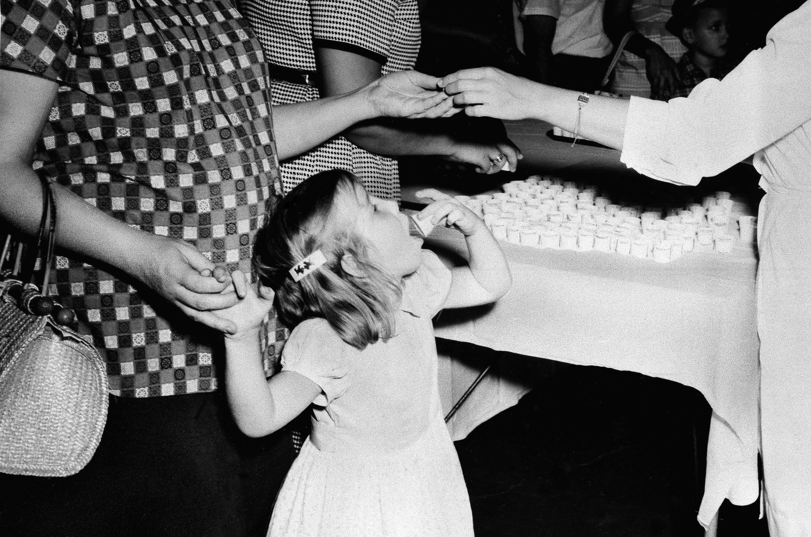 A young girl swallows a vaccine-coated sugar cube, standing beside several adults and a table covered in small paper cups.