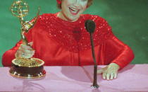 Woman in a red dress holding a golden Emmy trophy, smiling in front of a microphone.