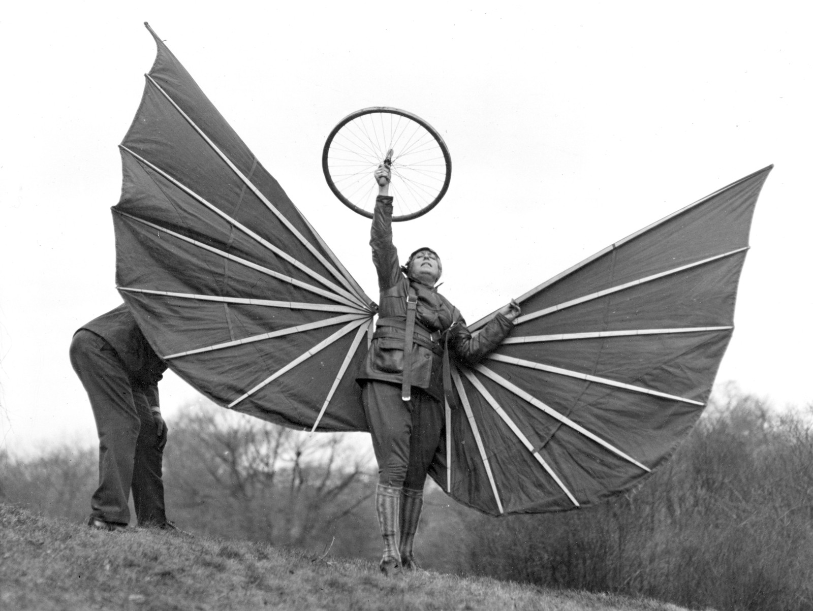 A person wearing an aviator costume, outfitted with fabric and wood wings, stands on a hill, holding a spoked wheel above their head.