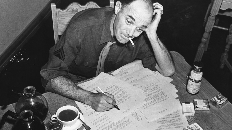Black-and-white photo of a man smoking a cigarette and filling out forms that are scattered on his desk with two pots of coffee and one cup of coffee next to the paperwork