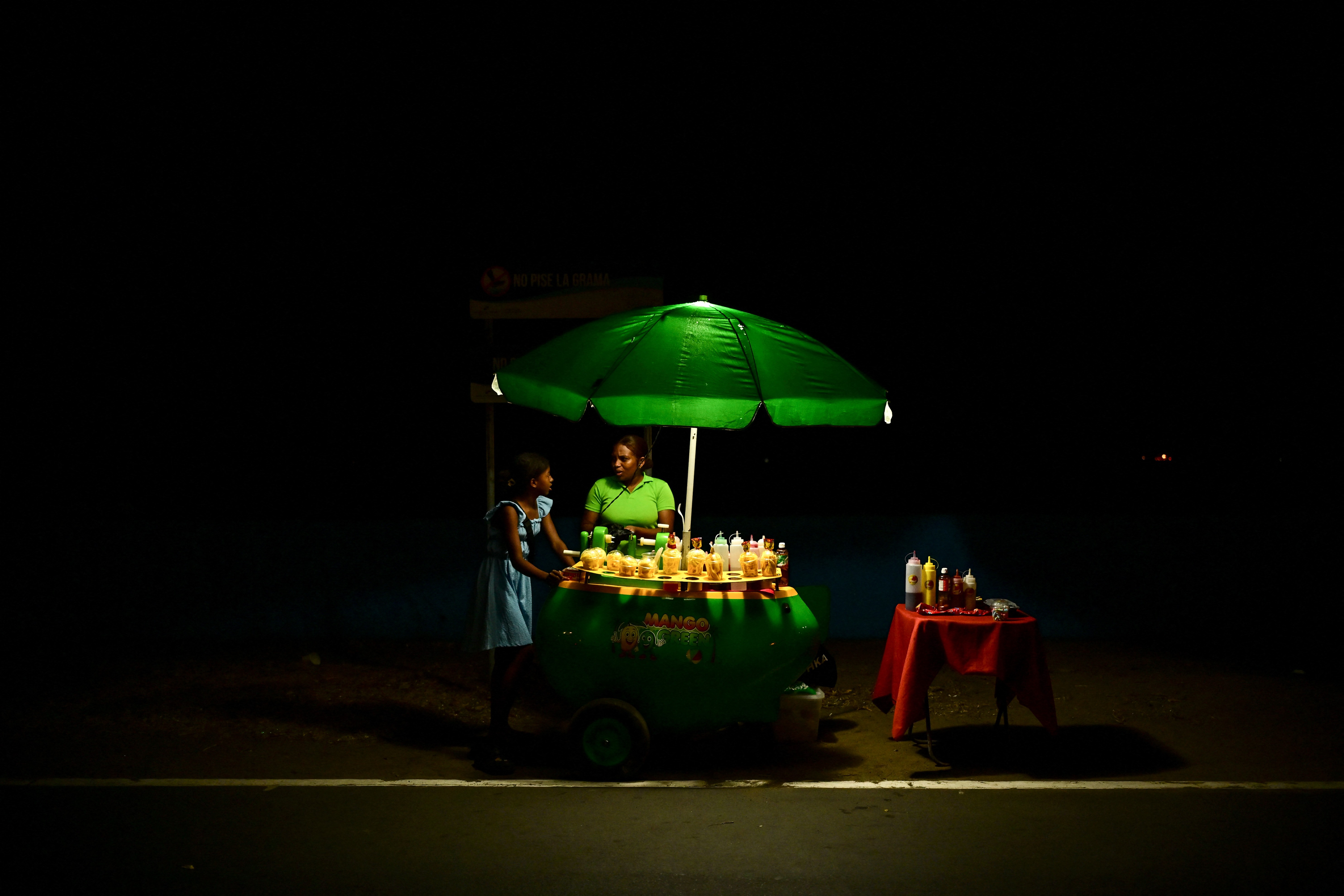 A woman sells mangoes from a street cart, seen at night.