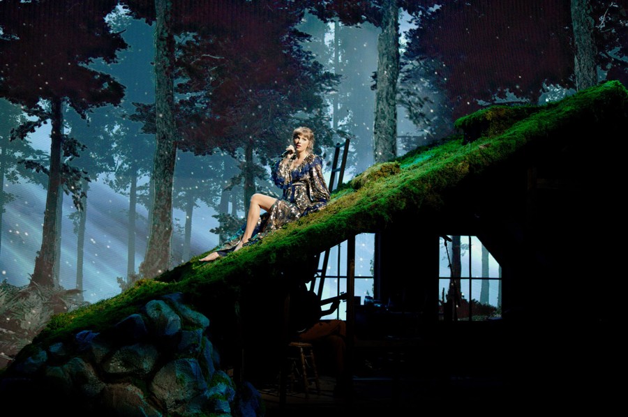 Performer Taylor swift sings while seated on the grassy roof of a cabin, built as a stage set.