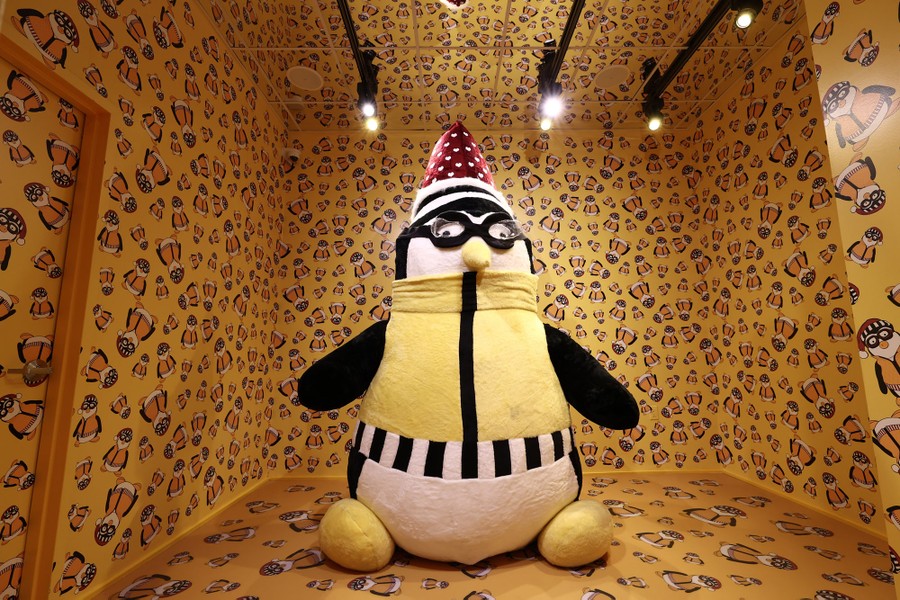 An enormous stuffed penguin sits in the middle of a room, with wallpaper on the walls covered in smaller images of the penguin.