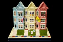 Sculpture of 3 three-story rowhouses, blue, white, red, with green grass and front steps. The middle house has an upside-down U.S. flag and Q-Anon flag, yard signs about J6, Trump flag in window, and other signs.