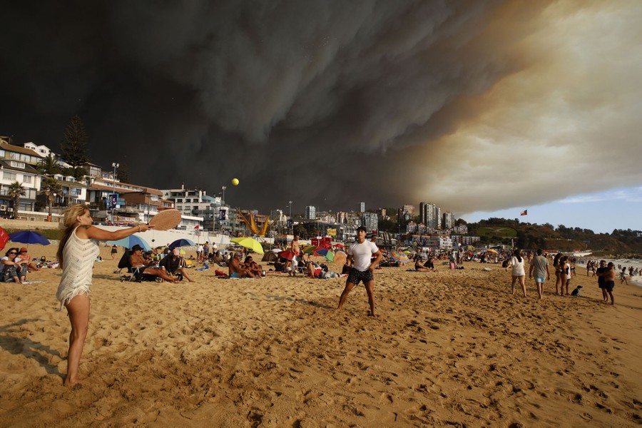 People walk and play on a beach as the sky above them fills with a massive black cloud of wildfire smoke in the sky above them.