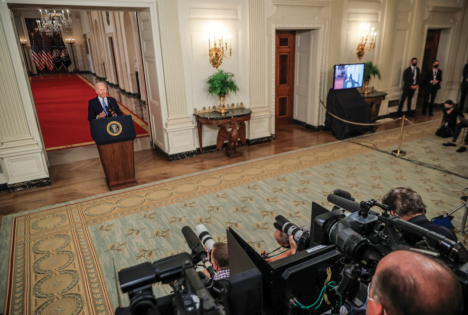 photo of President Biden speaking behind lectern with presidential seal, with hallway behind; the numerous cameras, microphones, and reporters recording him; and staff to the side near a television 