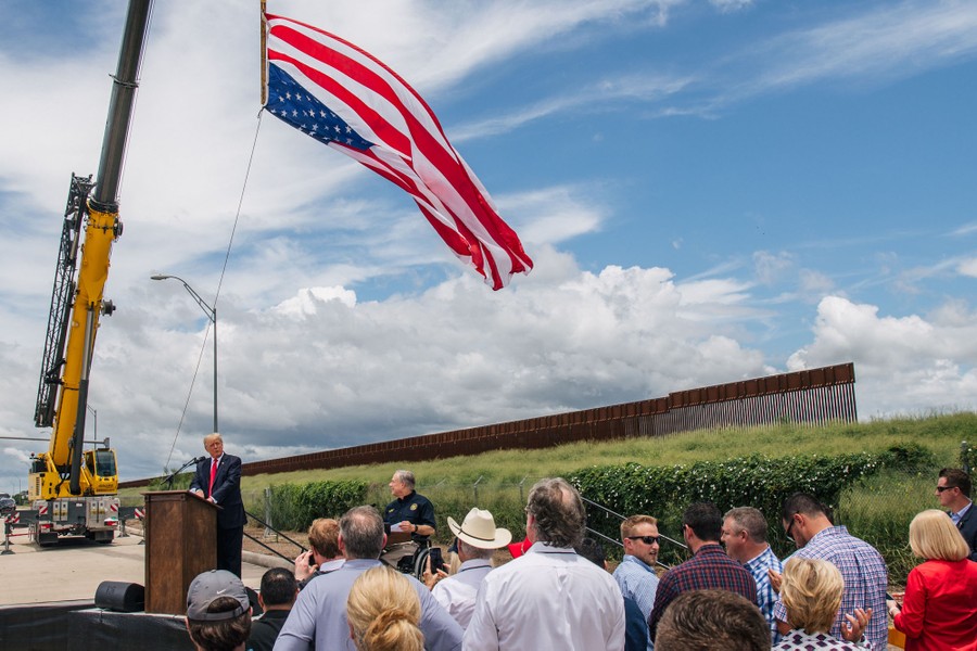 A small group of people can be seen looking up toward former President Trump at a podium, beneath a large American flag, in front of an unfinished section of border wall.
