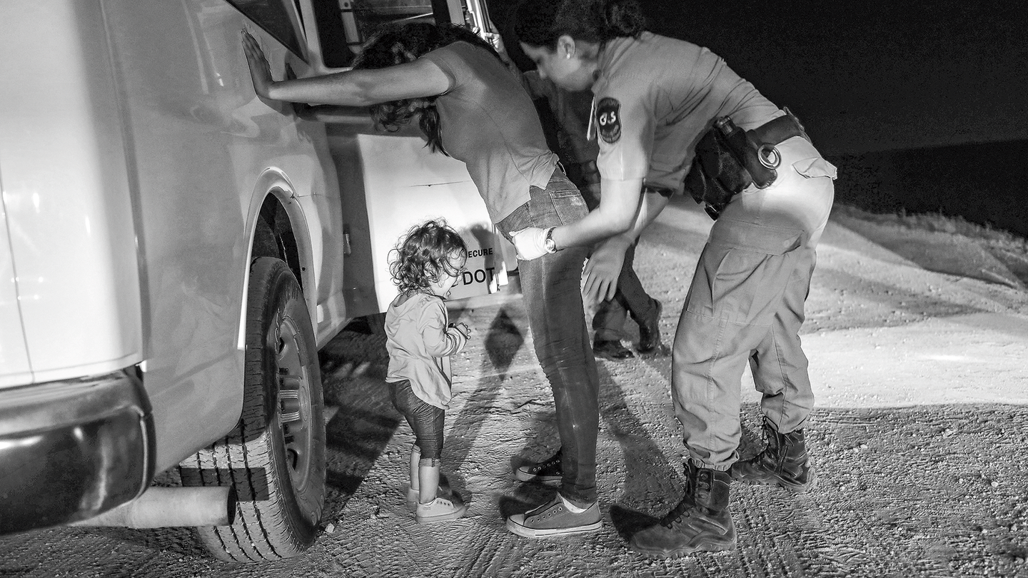 Una fotografía en blanco y negro de una mujer con una camiseta y jeans apoyada con ambas manos contra una camioneta blanca,mientras una mujer uniformada la inspecciona y una niña pequeña llora, por la noche.