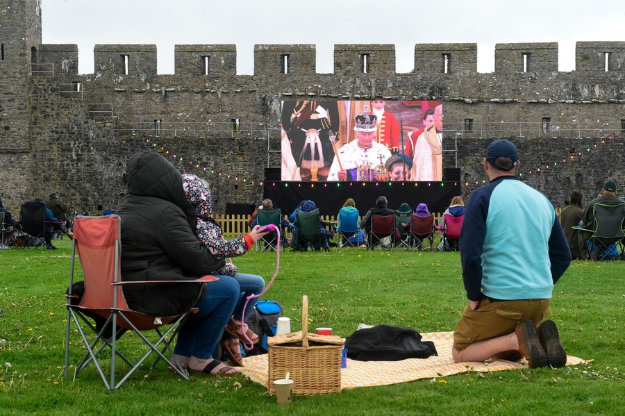 A small crowd watches the ceremony on a video screen set up against a castle wall.