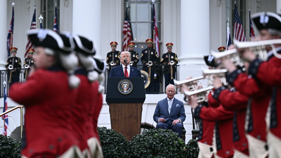 Photograph of President Trump standing at a podium with King Charles III seated beside him and the White House behind both of them. Soldiers wearing red uniforms play the trumpet and march in front of the leaders.