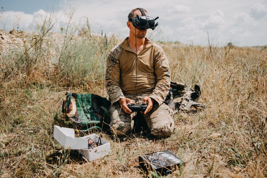 A soldier kneels in a field, wearing a headset, and operating controls for a remote drone.