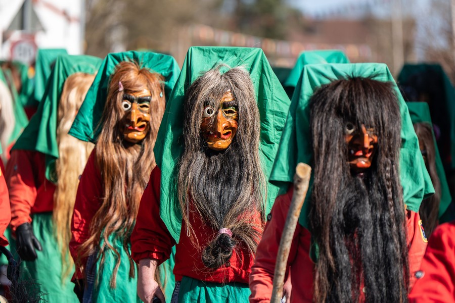 Performers wearing wooden masks with long hair march in a parade.