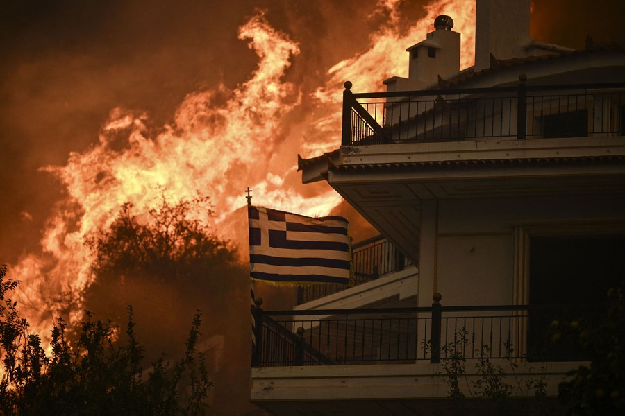 A Greek flag flies in the wind, mounted to a building's porch, with a roaring wildfire filling most of the background.