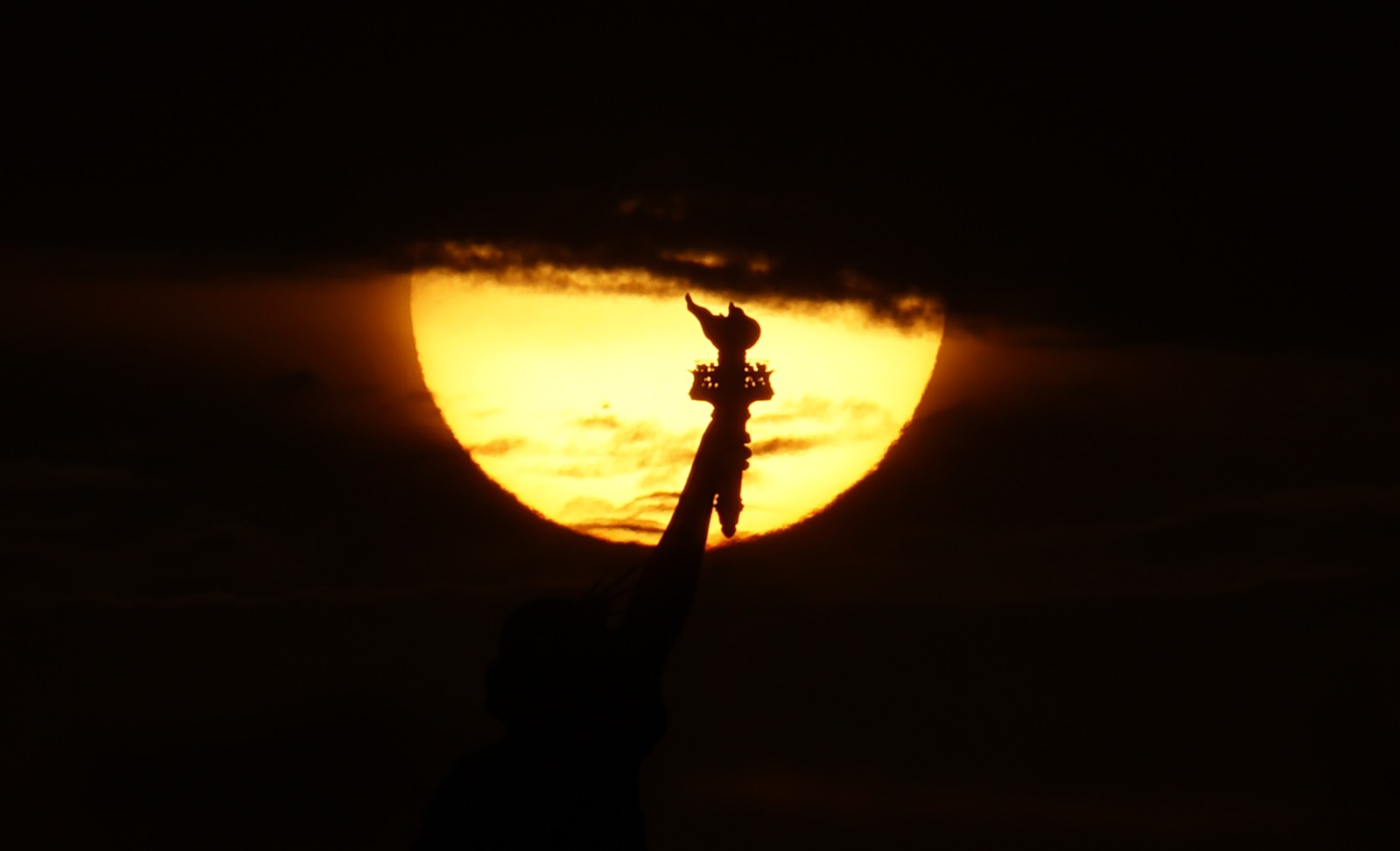 The sun rises behind the upraised arm and torch of the Statue of Liberty.