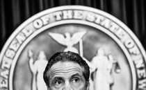 The top half of New York governor Andrew Cuomo's head is shown in front of the seal of the state of New York.