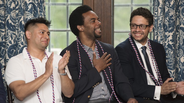 Three men sit with red and white graduation chords. One has his hand over his heart