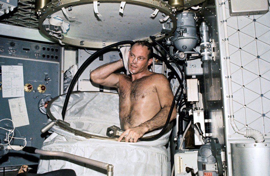 A man takes a shower on a space station inside a flexible shower curtain that has been partially lowered to show the mechanism.