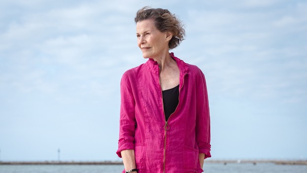 Woman in magenta top and black capri pants standing barefoot on beach with cloudy blue sky behind