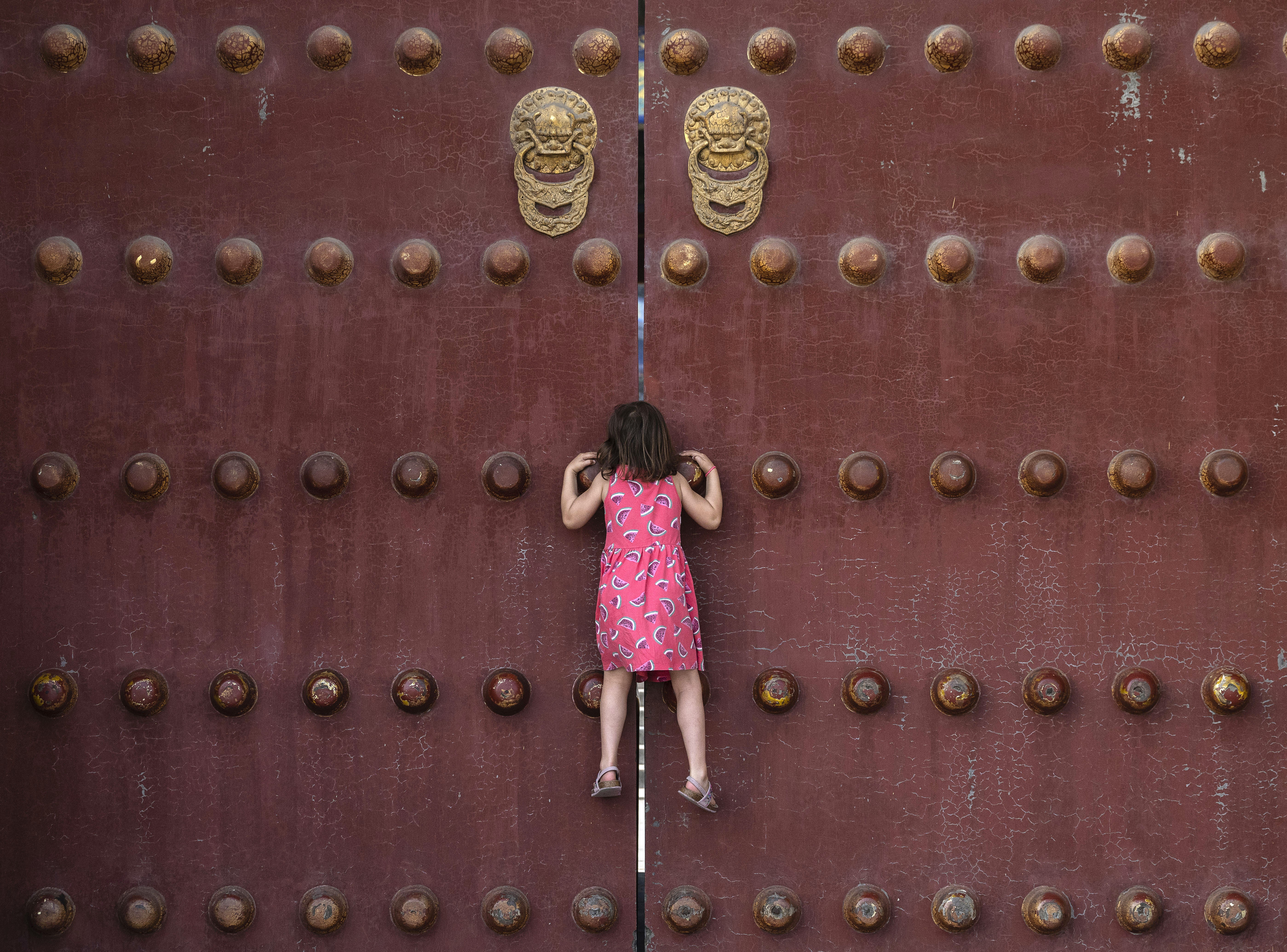 A young girl climbs to look through a gap of the large closed doors of a temple.