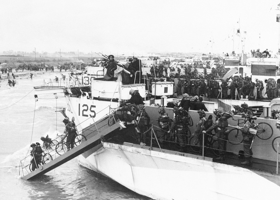 Soldiers carry bicycles as they disembark from several landing craft along a beach.