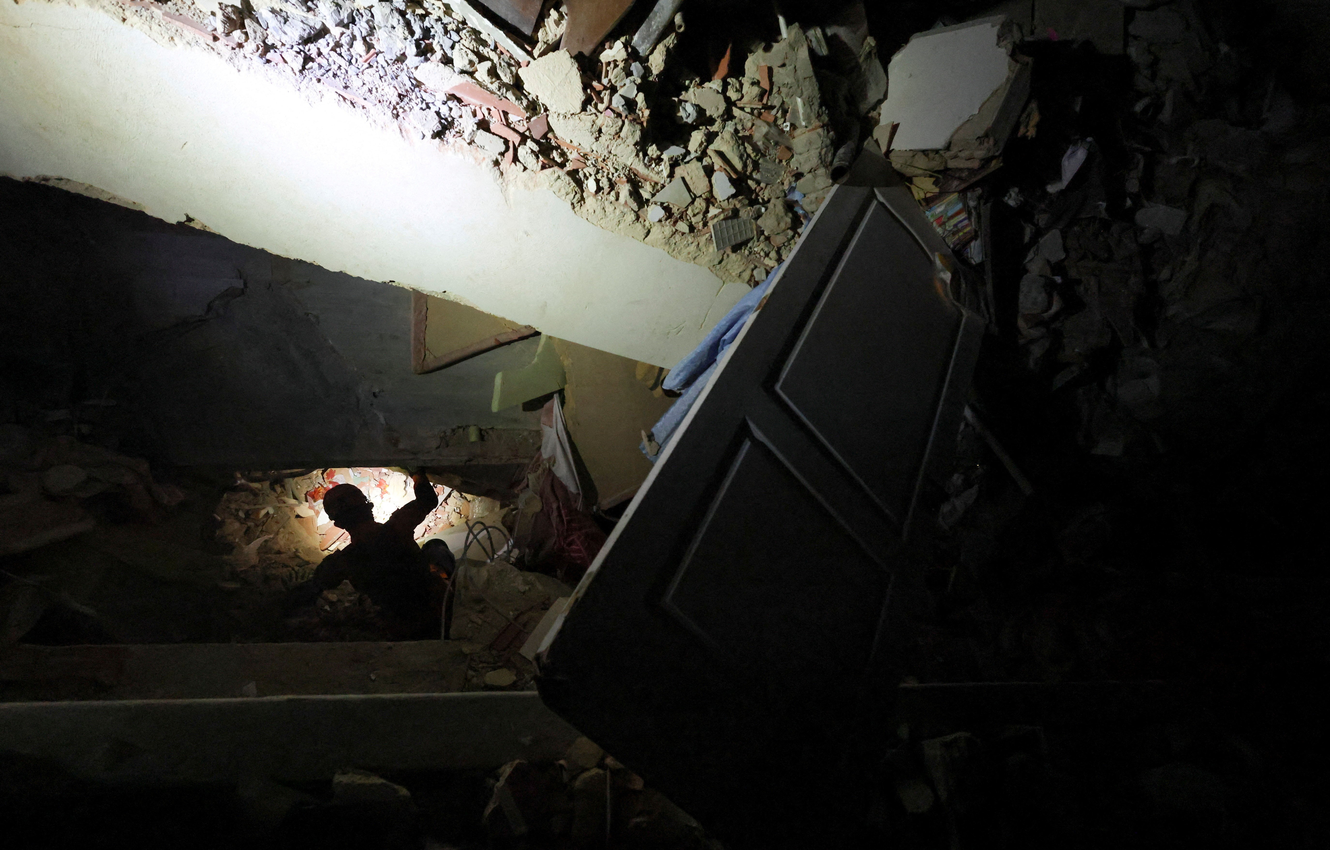 A rescue worker wearing a headlamp searches through the rubble of a large collapsed building in the dark.