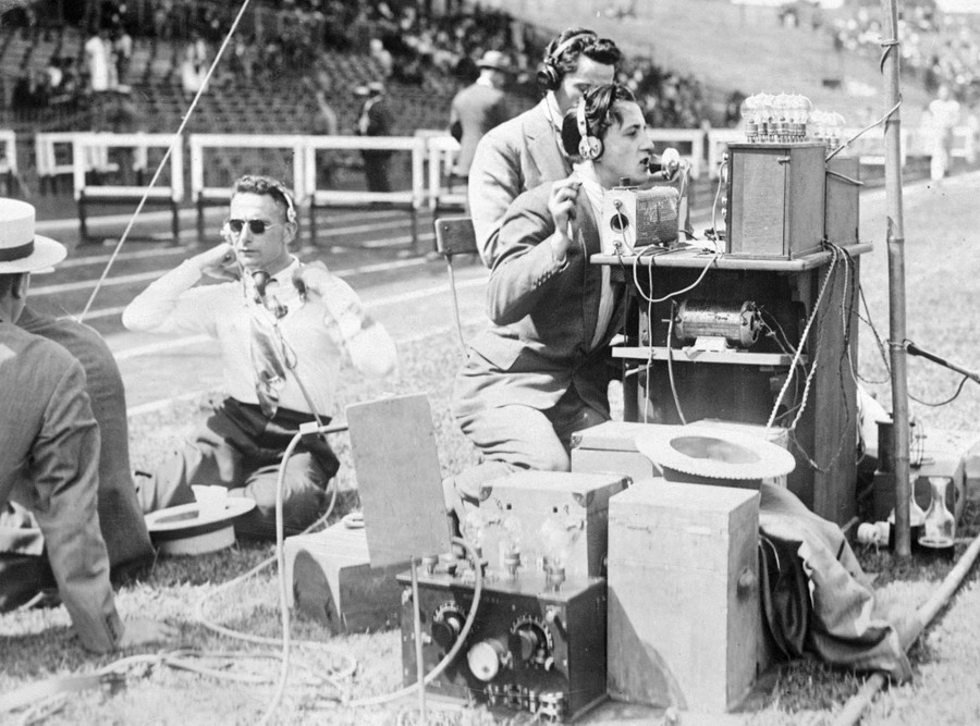 Several people use early radio equipment set up inside a race track in a stadium.