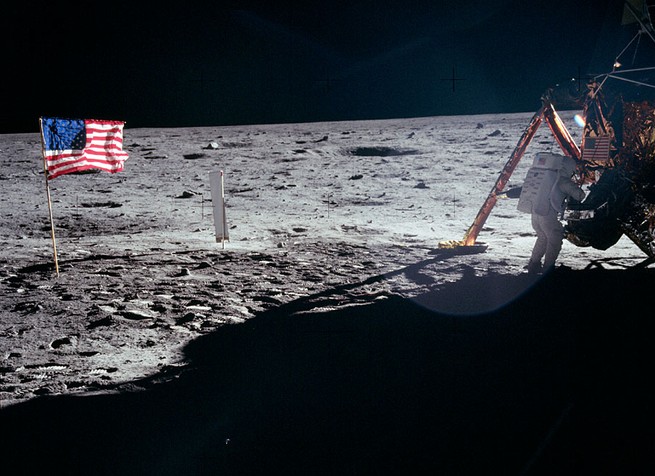 Neil Armstrong on the surface of the moon by the lunar module, with the American flag to his left.