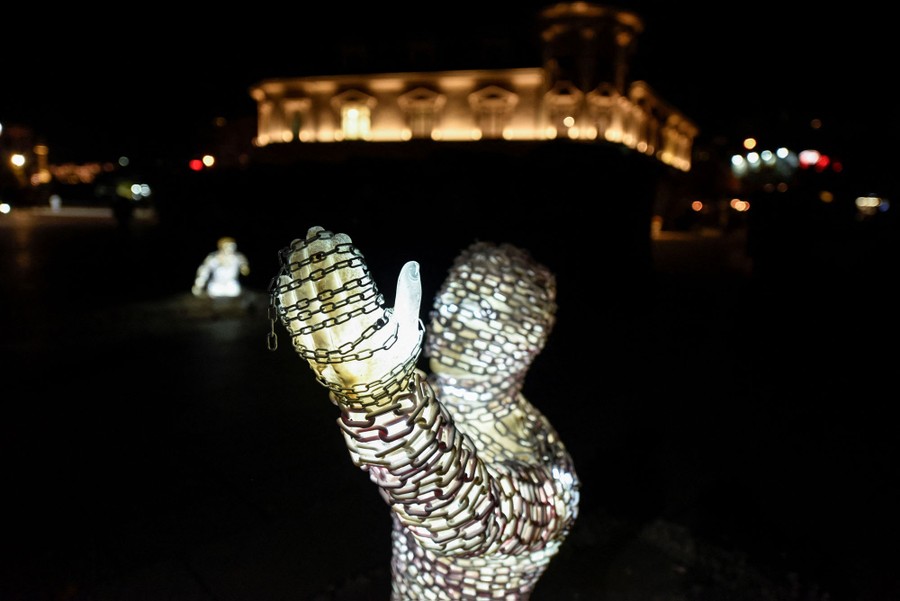 Illuminated statues, wrapped in chains, are seen in a city square at night.