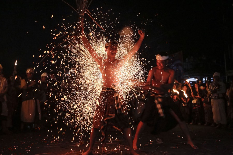 A person strikes another with burning coconut leaves, sending up a shower of sparks.
