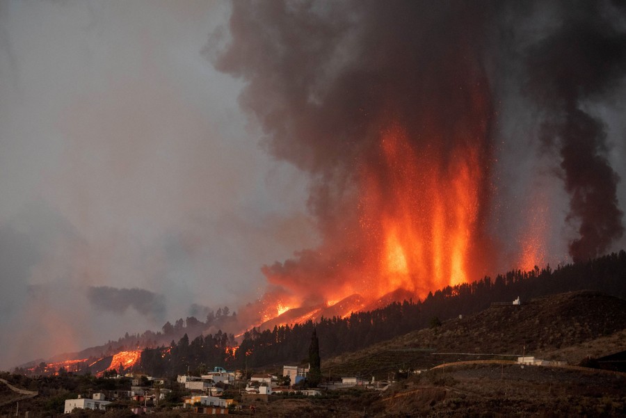 Lava and ash spew from the slope of a volcano in huge jets above several houses.