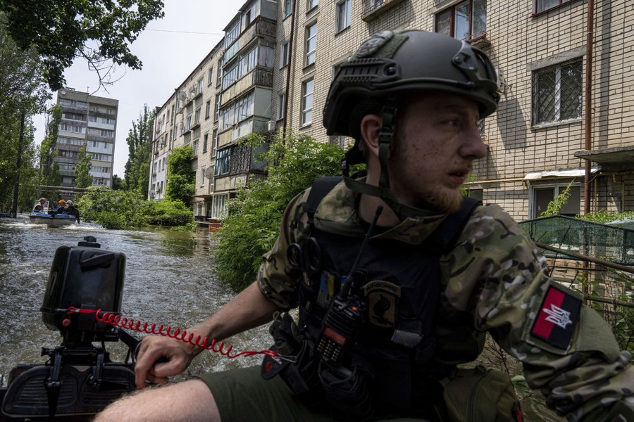 A soldier steers a boat through floodwater amid residential buildings.