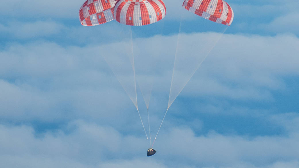 A gumdrop-shaped spacecraft, charred from the heat of reentry, coasts through blue sky beneath a trio of red-and-white parachutes.