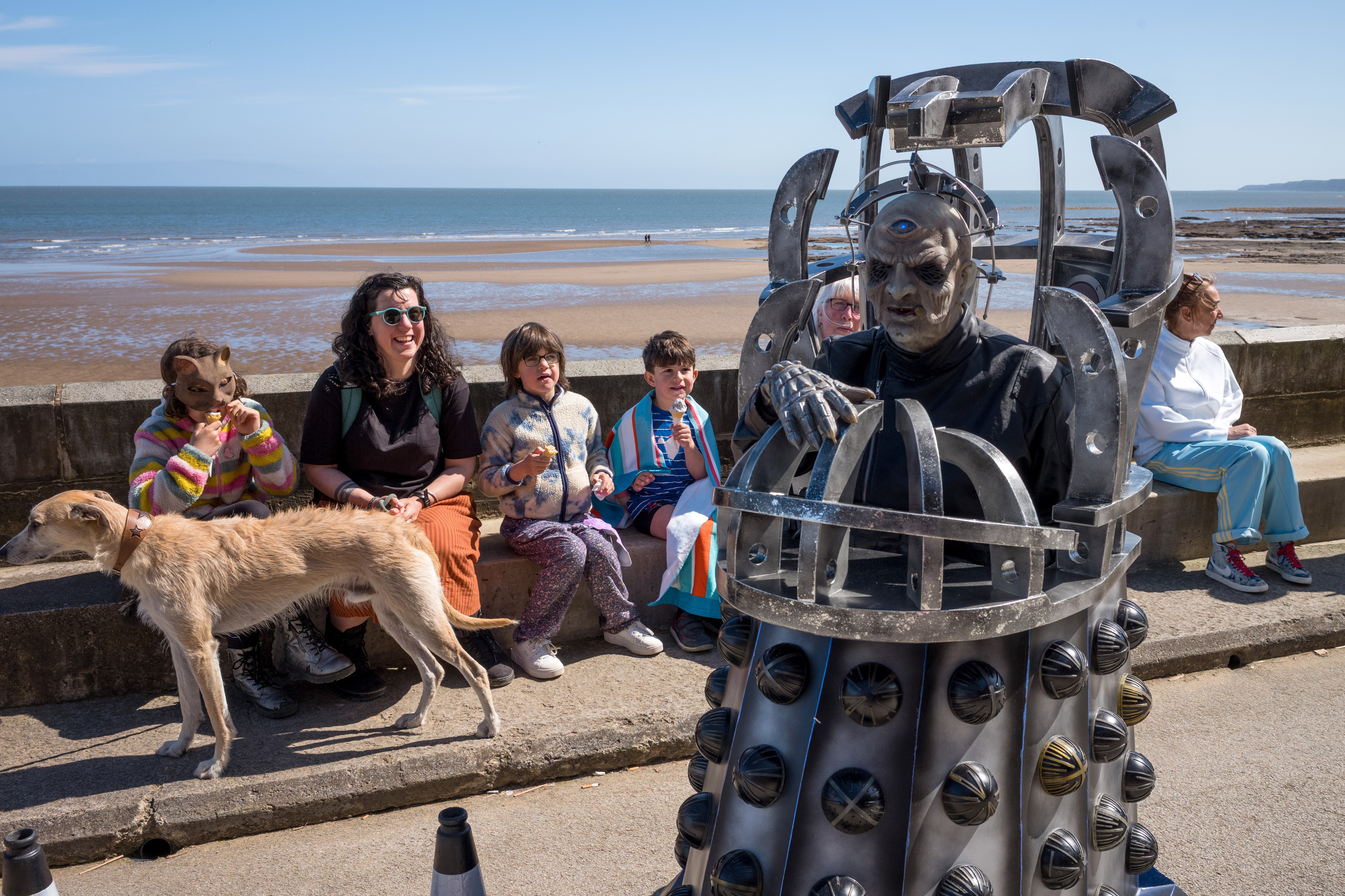 Visitors on a seafront bench watch a performer dressed in a costume based on a 'Doctor Who' villain.