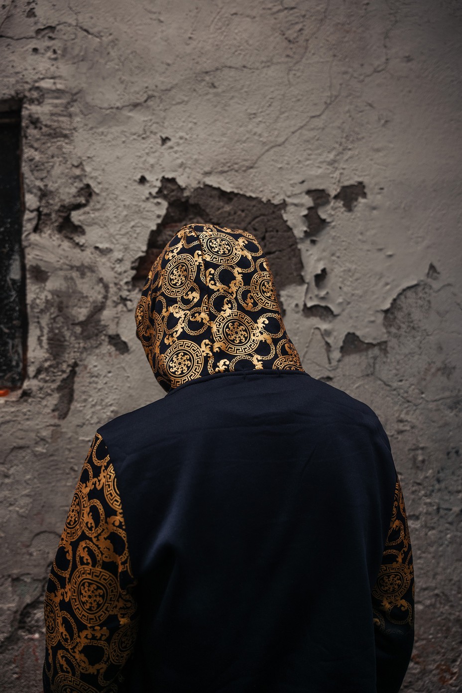 a man in a black and gold hoodie facing a white concrete wall