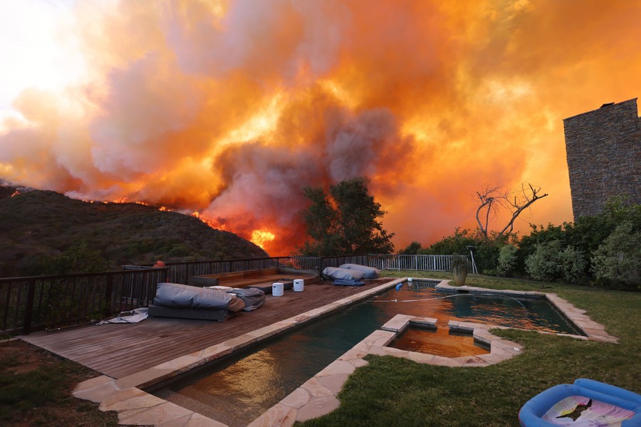 Smoke rises from a fire burning on a hillside in the distance behind the backyard of a house with a swimming pool.