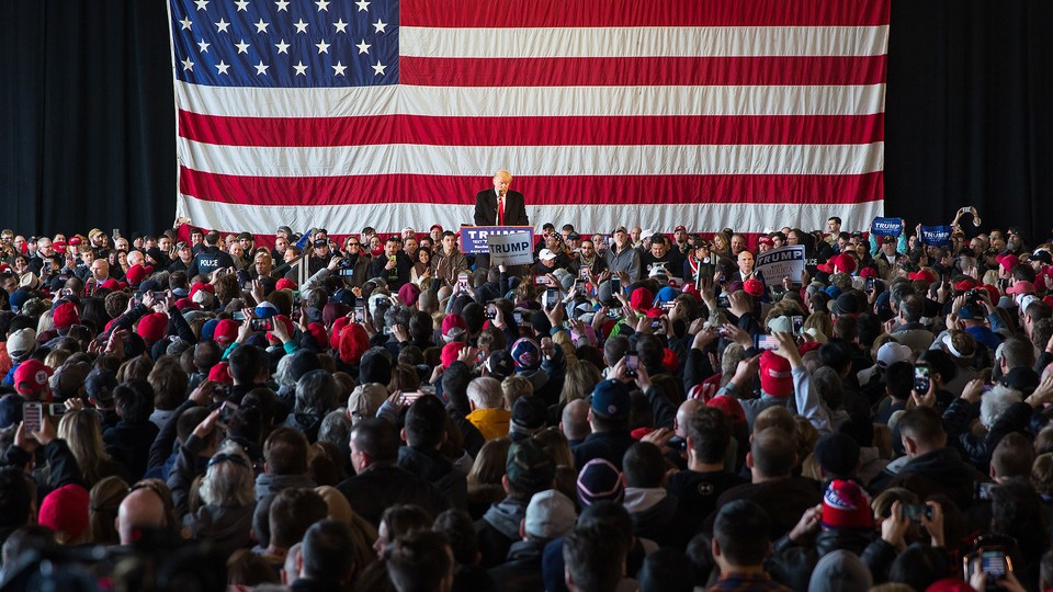 Donald Trump addresses a crowd of supporters with a large American flag behind him.