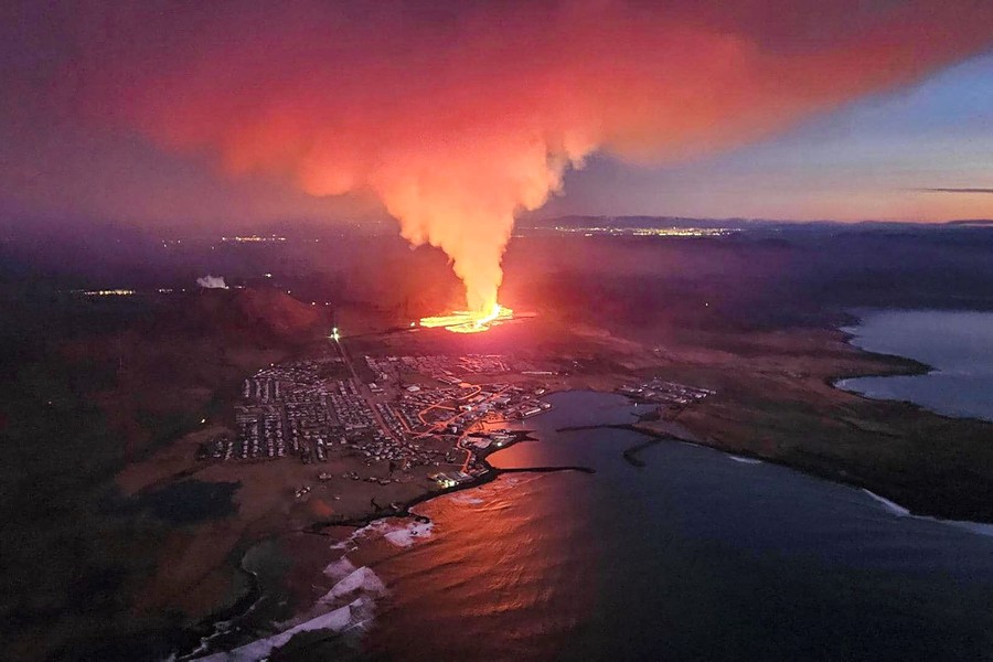 An aerial view of a coastal Icelandic town with an erupting volcanic fissure a short distance inland, just uphill from houses